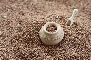 Background image of a large pile of buckwheat, in the middle of which lies a small jug and a wooden spatula for cereals photo