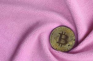 The golden bitcoin lies on a blanket made of soft and fluffy light pink fleece fabric with a large number of relief folds. The shape of the folds resembles a fan from a video card cooler photo