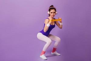 Slender girl involved in sports on a purple background. Woman in 80s style sports outfit squatting photo