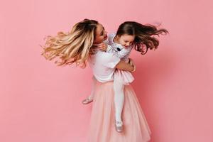 Blonde woman holds daughter in her arms and spins on pink background. Snapshot of curly mom and chi