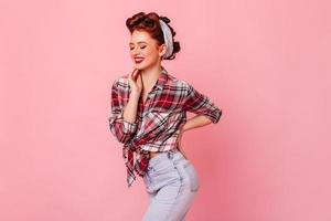 Graceful fit girl in checkered shirt standing on pink background. Studio shot of dancing pinup woma photo