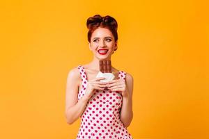 Good-humoured european girl eating chocolate. Pinup young woman in polka-dot dress smiling on yello photo