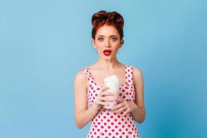 Surprised girl with red lips drinking milkshake. Studio shot of emotional young lady in polka-dot d
