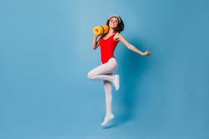 Girl in tight spots suit happily jumps on blue background. 80s woman carries her yellow fitness mat