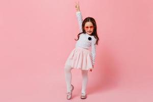 Stylish little girl in fluffy skirt, polka dot blouse and heart-shaped sunglasses shows peace sign photo