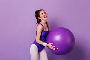 Beautiful girl in purple bodysuit and leggings does exercises with huge fitball on isolated backgro