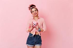 Charming woman with red lips in pin-up style outfit posing with pink mini camera on isolated backgr photo