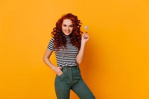 Attractive green-eyed redhead girl in cropped top and jeans smiling and posing with lollipop in ora