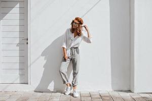 Stylish girl in gray pants and white cotton blouse posing near white wall. Photo of woman in cap an