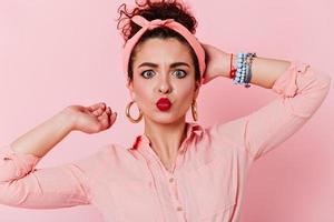 Pin-up girl posing on pink background. Close-up shot of blue-eyed girl with red lipstick in headban