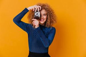 Positive woman in striped blue sweater takes photo on retro camera on yellow background