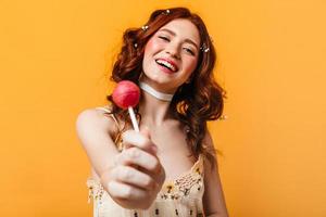 Sweet girl with red curls smiles and holds lollipop on orange background
