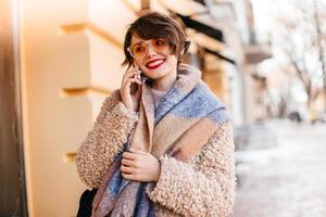 Wonderful young woman talking on phone on street. Outdoor shot of trendy girl in coat and scarf. photo