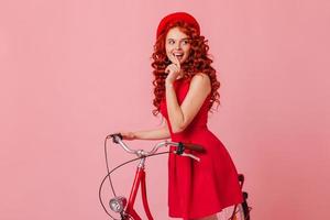 Charming coquettish woman in great mood looks slyly to side, posing with bicycle on pink background photo