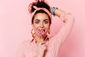 Surprised blue-eyed brunette girl in pink pin-up outfit eats lollipop on isolated background photo