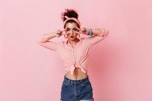 Stylish pin-up girl with pink headband showing peace signs. Portrait of woman in pink blouse and sh