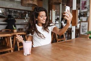 Young lady in cafe with wooden furniture makes selfie. Charming woman in white top took strawberry photo