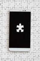 A modern big smartphone with several puzzle elements on the touch screen lies on a white jigsaw puzzle in an assembled state photo