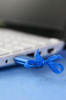 A brilliant blue USB flash drive with a blue bow is connected to a white laptop, which lies on a blanket of soft and fluffy light blue fleece fabric. Classic female design for a memory card photo