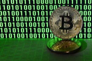 A pair of bitcoins lies on a cardboard surface on the background of a monitor depicting a binary code of bright green zeros and one units on a black background. Low key lighting photo