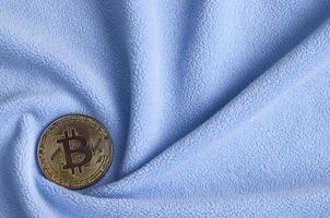 The golden bitcoin lies on a blanket made of soft and fluffy light blue fleece fabric with a large number of relief folds. The shape of the folds resembles a fan from a video card cooler photo