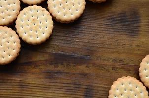 A round sandwich cookie with coconut filling lies in large quantities on a brown wooden surface. Photo of edible treats on a wooden background with copy space