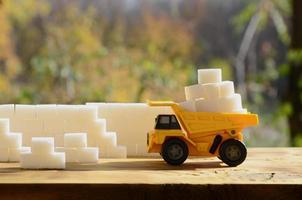 A small yellow toy truck is loaded with white sugar cubes near the sugar wall. A car on a wooden surface against a background of autumn forest. Extraction and transportation of sugar photo