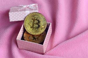 The golden bitcoin lies in a small pink gift box with a small bow on a blanket made of soft and fluffy light pink fleece fabric with a large number of relief folds photo
