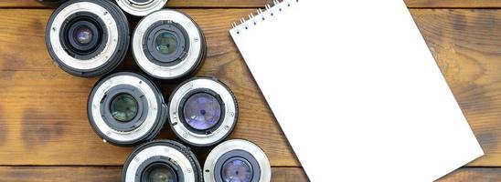 Several photographic lenses and white notebook lie on a brown wooden background. Space for text photo