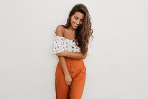 Stylish long-haired curly woman in orange trousers and white cropped top smiling shyly on isolated
