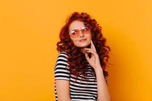 retrato de primer plano de una adorable niña pensativa con cabello rojo ondulado que usa camiseta y anteojos en forma de o