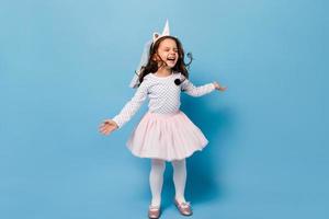 Happy female kid in fluffy skirt with unicorn horn laughs and jumps on blue background photo