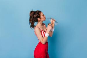 Dark-haired girl with massive earrings in red top and trousers drinking water on blue background photo