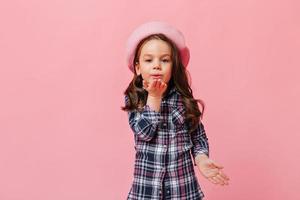 Female curly kid in blue shirt and beret in French style blowing kiss on pink background photo