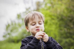 A happy boy on a spring day in the garden blows on white dandelions, fluff flies off him. The concept of outdoor recreation in childhood. Portrait of a cute boy. photo