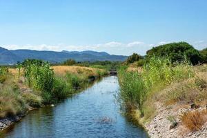 view of a water channel among grass and bushes at a clear sunny day with a mountains on a background photo