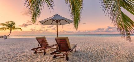 Beautiful tropical sunset beach. Romantic couple goals, two sun beds, umbrella under palm tree leaves. Panoramic coast landscape, love and relaxation banner. Colorful dream travel sea sand sky island