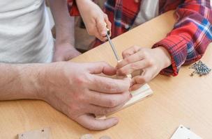 Happy father's day and childhood concept. Close-up of a father and a boy son in glasses work with hand tools, using a screwdriver, assembling a wooden house constructor at the table. photo