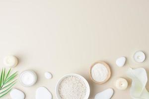 Spa background with bowl of salt, skin care products, white stones, candles, palm leaf and white flower on pastel beige. Flat lay, copy space photo