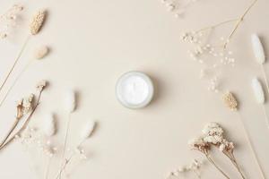 Jar of cosmetic cream with flowers on pastel beige background. Flat lay photo