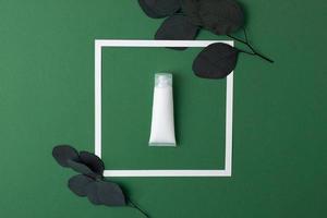 White tube of cosmetic cream with white frame and eucalyptus on green background. Flat lay photo