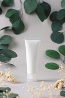 White tube of cosmetic cream with flowers and green leaves on grey background. Close up photo