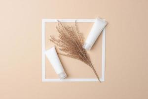 White tubes of cosmetic cream with white frame and flowers on pastel beige background. Flat lay, copy space photo