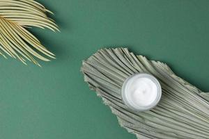 Jar of cosmetic cream with leaf palm branch on dark olive background. Flat lay, copy space photo