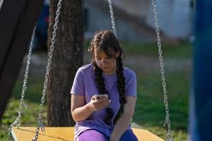 a teenager girl swings on a swing with a phone in her hands, communicates in social networks or learns online photo