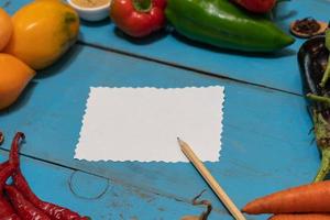 Vegetables are laid out around a sheet of paper and a pencil. Empty space for text. Vegetables, empty blank for recipe on a blue background. photo