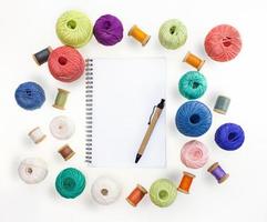 Multicolored balls and bobbins of woolen yarn, wooden thread sleeves on white isolated background. Notepad with pen. Needlework, handmade. view from above. Isolated. Copy space photo