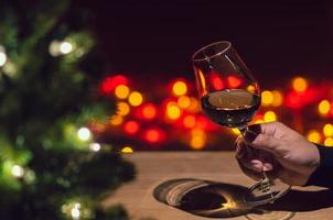 Hand toasting a glass of Rose wine on wooden table with Christmas tree and colorful bokeh light background.