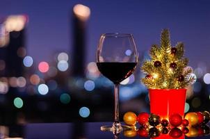 A glass of red wine that have Christmas tree and bauble ornaments with lights on colorful city bokeh light background. photo