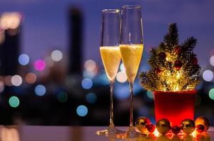 dos copas de vino espumoso con árbol de Navidad y adornos de adorno puestos en la mesa con un colorido fondo de luces de la ciudad. foto